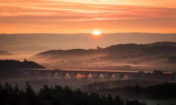 Das Vogtland bei Sonnenaufgang: Nebel über bewaldeten Hügeln und einer Eisenbahnbrücke – die Heimatregion von Autohaus Voitel in Plauen
